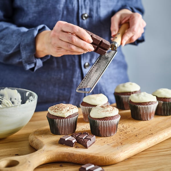 Person grates chocolate over frosted cupcakes on wooden board. Bowl with icing nearby. Zester with wooden handle visible.