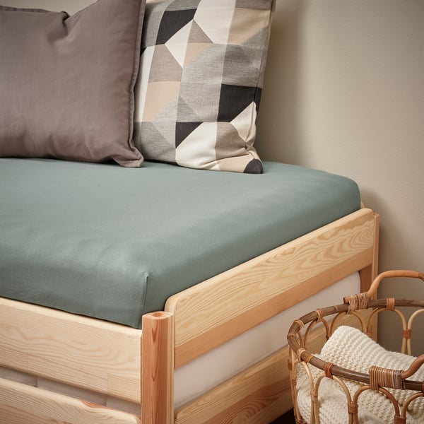 Bed with teal VÅRVIAL sheet, two small pillows, beige wall, basket with blanket.