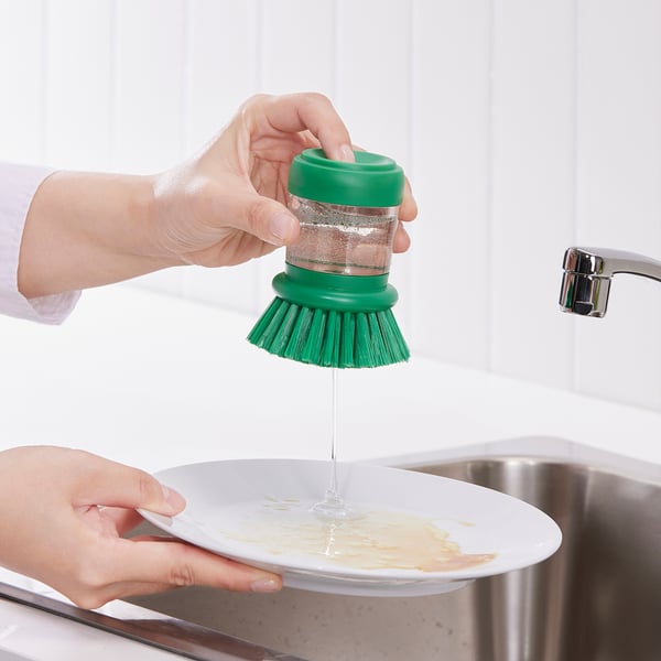 Hand holds green brush releasing soap onto dirty plate. Metal sink and tap nearby.