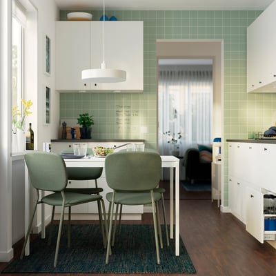 Modern kitchen with small white extendable dining table, green chairs, and backsplash tiles. Dark rug contrasts wood floor.