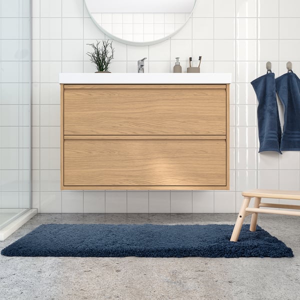 Modern bathroom with large wooden cabinet, circular mirror, blue towels & plush ALMTJÄRN blue bathmat on speckled floor.