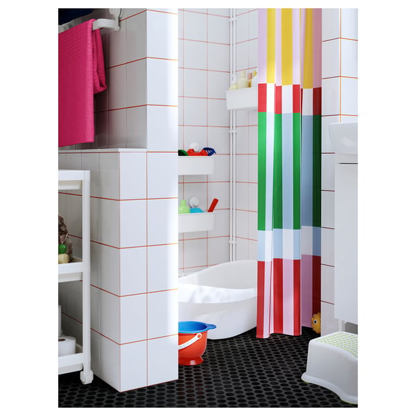 Kids bathroom with colourful BANJANTRÄD curtain, white tiles, and red bucket. Child-friendly design with bright colours.