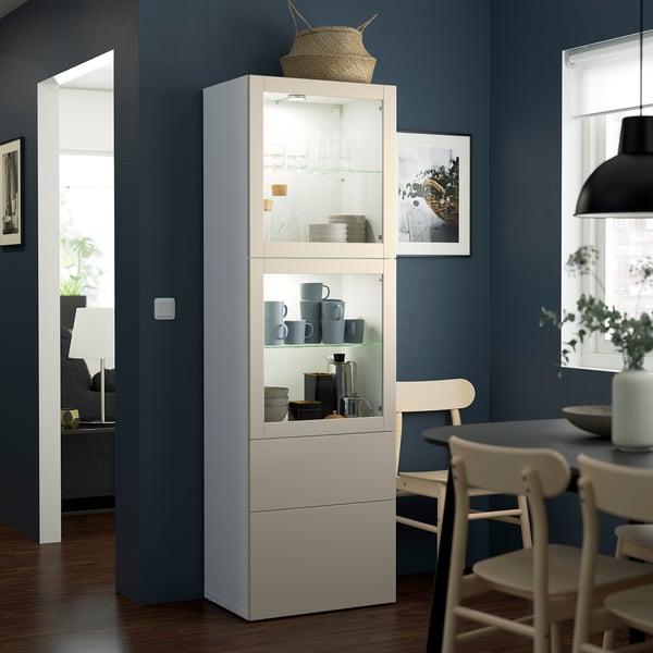 White BESTÅ cabinet with glass doors, blue mugs, near dining table in dark room.