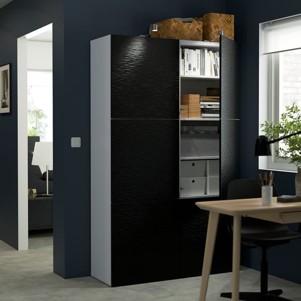 A tall black storage cabinet with one glass door revealing books and storage boxes. The cabinet is next to a table with a chair and a window.