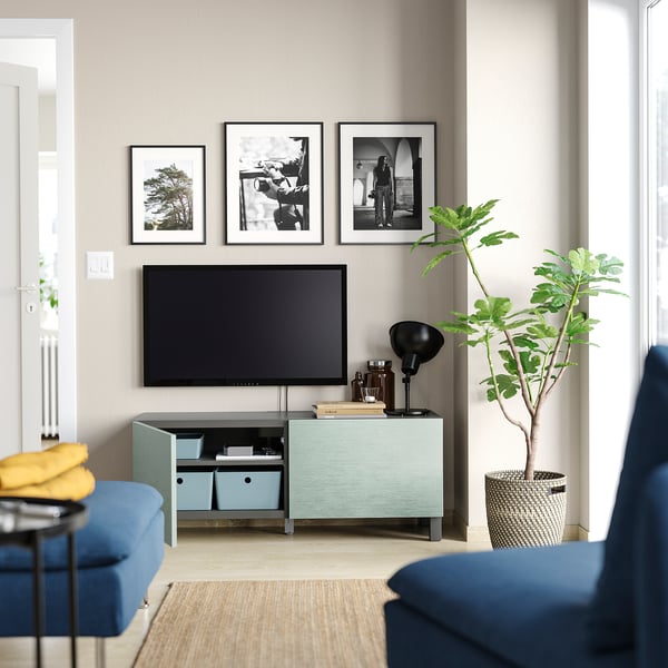 Modern living room with BESTÅ tv bench in white and blue. Flat-screen tv, framed photos, plant, and blue sofa.