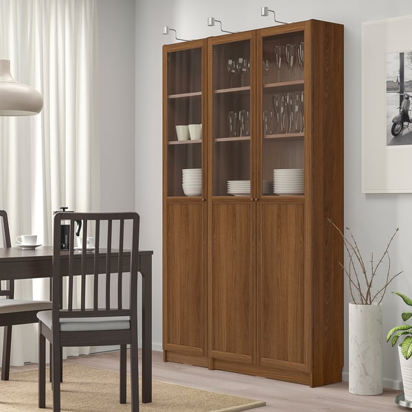 Large wooden BILLY bookcase in brown with glass doors, holding dishes & glasses, beside black chairs & dining table, white wall & curtain backdrop.