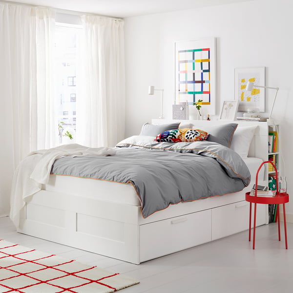 Minimalist white bed with grey bedding and colourful pillows, red rug and side table, bright room with abstract artwork.
