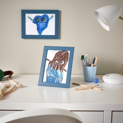 Childs desk with two drawings: blue monster, brown-haired figure. Art supplies, lamp.