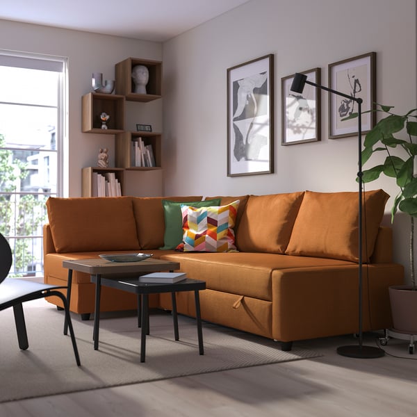 Modern orange FRIHETEN sofa bed, bright cushions, spacious, against wall with art, near window and plant.