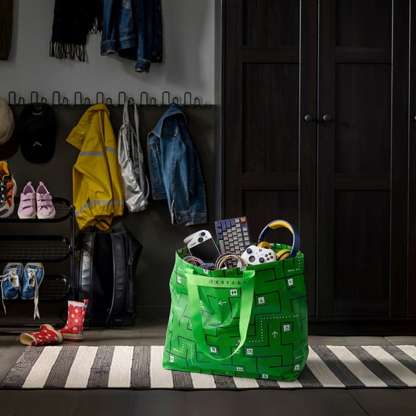 Bright green GÖTEHULT bag on striped rug, holding electronics, near entrance filled with shoes & coats.