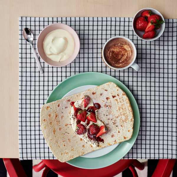 A meal setup on a chequered tablecloth featuring a folded crepe with berries, a cup of coffee, a bowl of cream, and a bowl of strawberries.