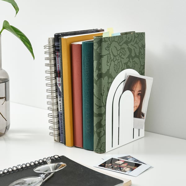 Bookshelf with white HÖSSLA bookends holding colourful books, plant, and photos.