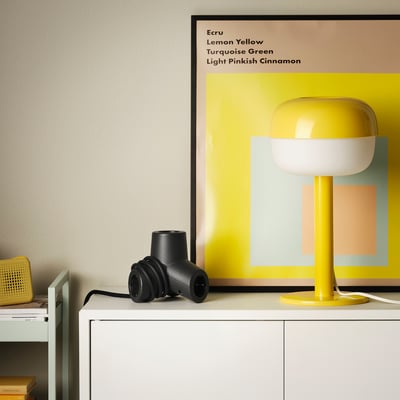 Black HORNFELS 4-way extension on a white cabinet, smaller than a yellow lamp, powers multiple devices with a unique, fun design.