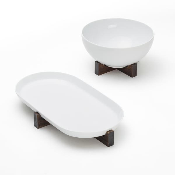 Two white ceramic bowls on dark wood stands. One is round, the other oval, both elevated for display. Contrast highlights elegance.
