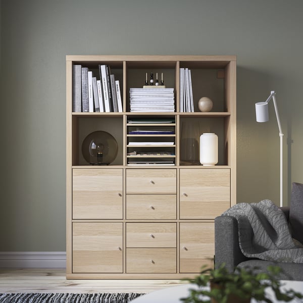 KALLAX light wood shelf unit with books, decor, and drawers against wall, grey sofa right.