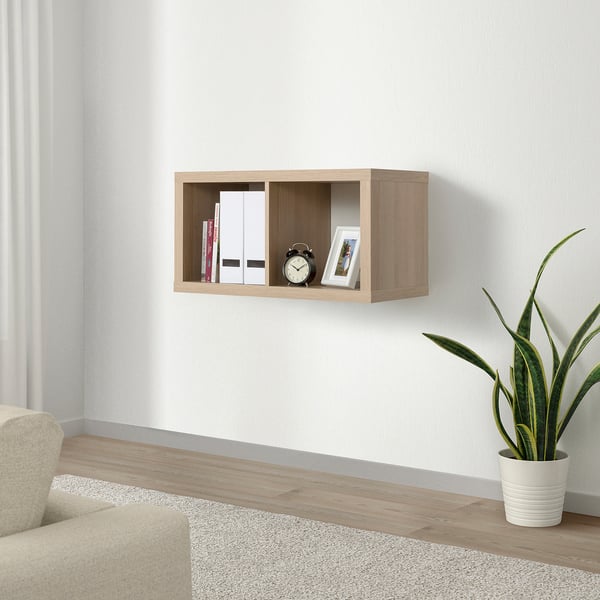 Floating KALLAX shelf in light brown holding books, clock, and framed photo. Next to green plant, light-colored couch, and grey rug.