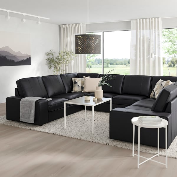 Modern living room with l-shaped black KIVIK leather sofa, large windows, white decor, cosy pillows, and nature view.