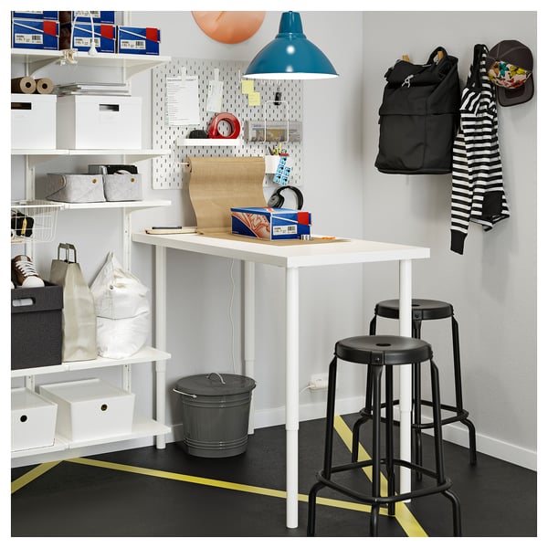 Well-organized workspace with white desk, two black stools, grey bin, shelves, and pegboard with various items.