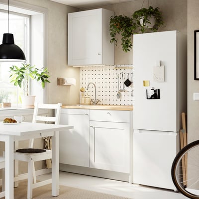 KNOXHULT compact white kitchen with sink, wooden top, white cabinets, black-cushioned chair, hanging plants, modern lamp, perfect for small spaces.