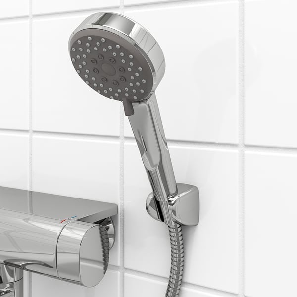 A chrome hand shower with holder, featuring multiple spray holes, mounted on a tiled wall. It includes a coiled hose and wall fittings.