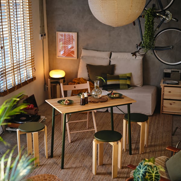 Small KOMPISHÄNG stools in birch & black by table, under hanging light, near cosy sofa & bicycle.
