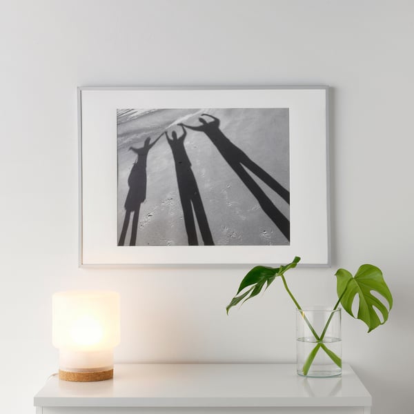 A white picture frame a black-and-white shadows on a white shelf. Beside it, a small lit lamp and a glass vase with a green plant.
