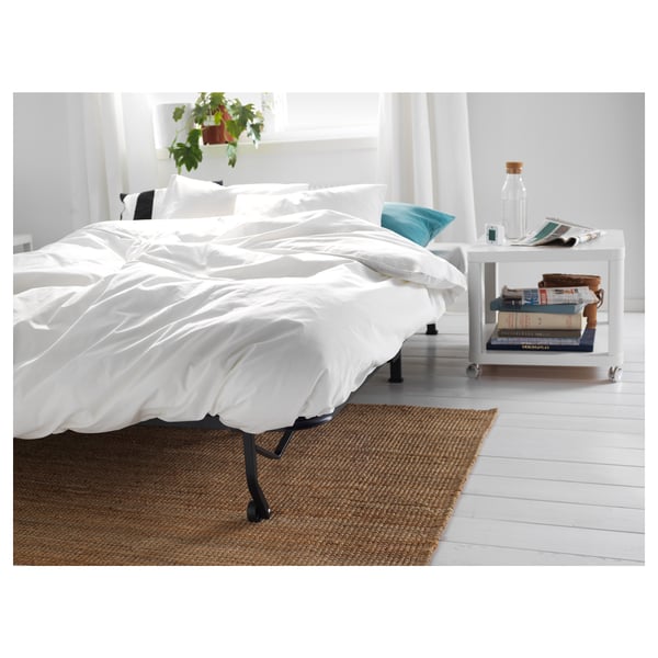 Minimalist white bed with black legs, crisp linens, small bedside table, books, bottle, plant, white floors, jute rug.