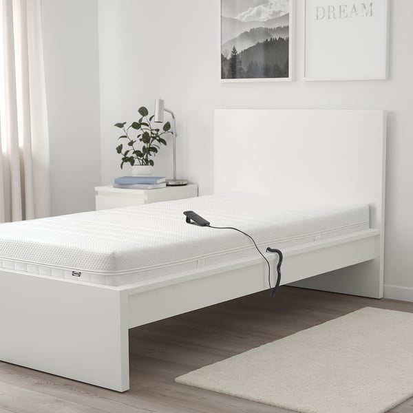 White MAJSKÄR foam mattress on bed with remote. Bigger than bedside table and rug, fits bed frame. Plant and lamp on bedside table.