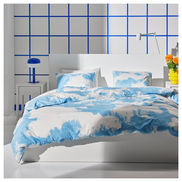 A MALM bed with blue-white cloud duvet, white headboard. Bold grid wall, bedside tables with lamp & flowers.