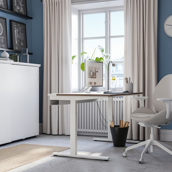 Modern home office setup. White MITTZON height-adjustable desk by window, wider than chair. Grey curtains, blue walls, plant & decor.