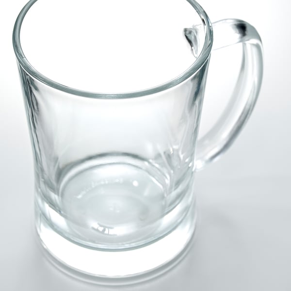 A clear, sturdy glass mug with a wide handle sits empty on a reflective surface, ideal for serving beer with a full head.