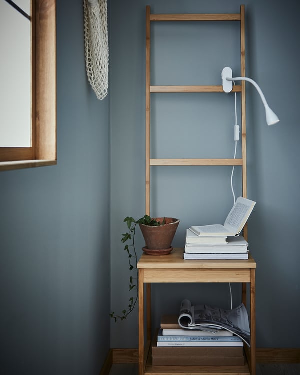 A small, slim, white NÄVLINGE clamp lamp attached to a wooden ladder shelf, illuminating a book on a wooden table against a blue wall.