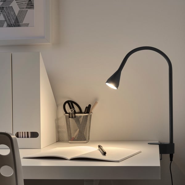 Black clamp spotlight on desk, illuminating book. Adjustable. Pen holder nearby.