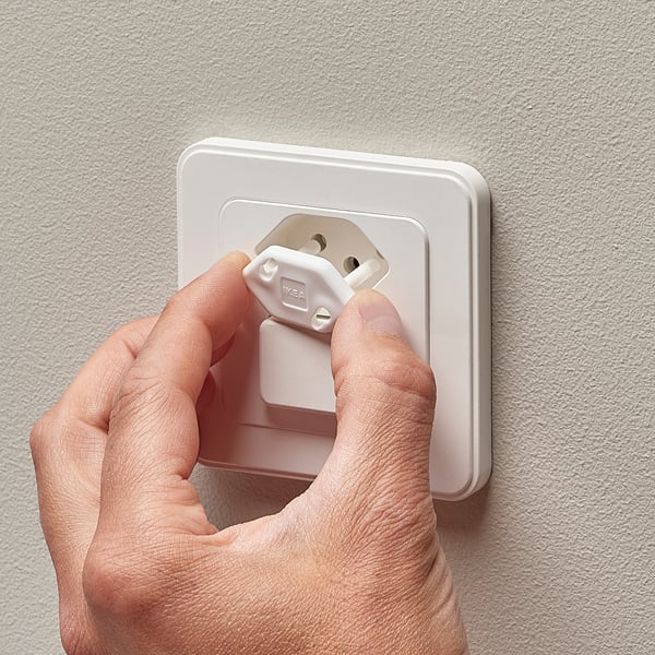 A hand inserts a white safety plug into a wall socket, demonstrating a childproofing method.
