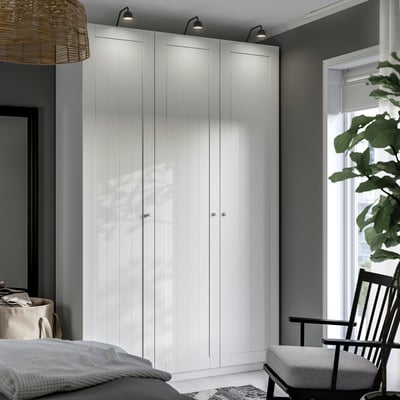 Modern bedroom with tall white PAX wardrobe, lights, rug, chair, bed, linens basket, and large plant.