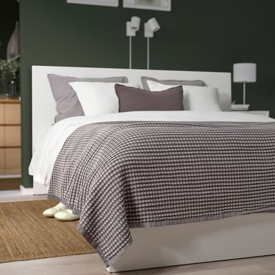 A grey cotton PELARTUJA bedspread with waffle weave on a double bed, two pillows, bedside table with lamp, and dresser with plant.