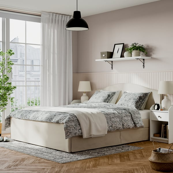 Beige RAMNEFJÄLL bed with headboard, flanked by nightstands. Patterned bedding, grey throw, plants on shelf, sheer curtains, pendant lamp, rug.