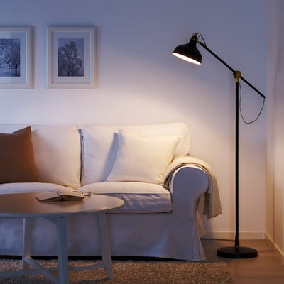 Cosy living room with white sofa, RANARP black floor lamp, brown rug, coffee table, and framed art. Lamp stands tall beside the couch.