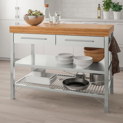 IKEA RIMFORSA grey kitchen island with bamboo top, glass shelves, and stainless steel accents, holding plates, bowls, and a frying pan.