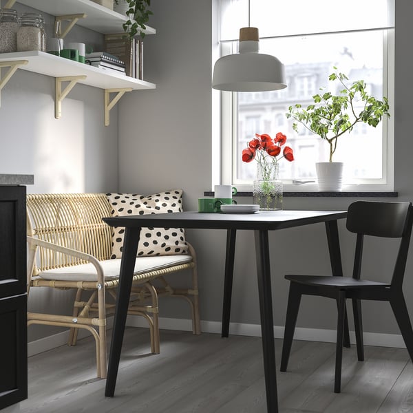 Natural rattan SALNÖ bench under white table, flanked by black chairs and shelves, with a bright window and plants.