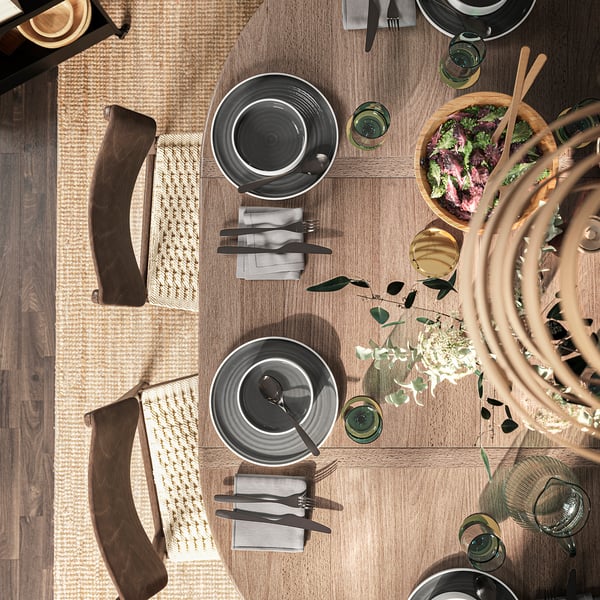 Top view of a modern, brown-stained, circular dining set, SKANSNÄS, with four dark grey plates, cutlery, glasses, and a bowl of salad.