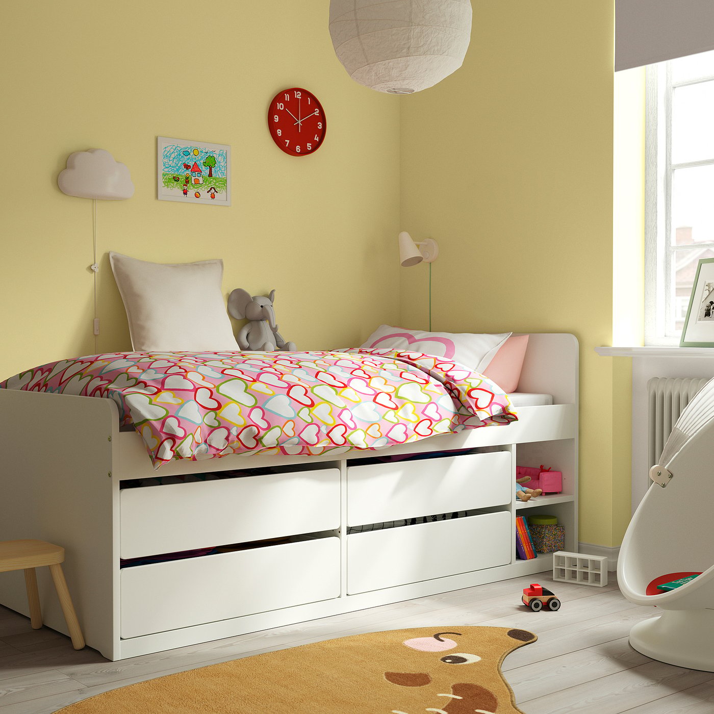 Bright yellow kids room with white SLÄKT storage bed, red clock, two lamps, stuffed elephant, colourful rug, and toys. Bed is central and large.