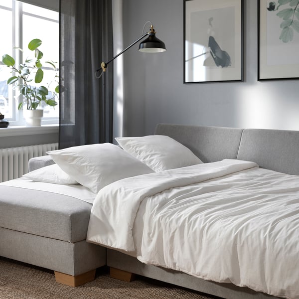 Modern sofa bed with white bedding, pillows, and plant beside a large window with curtains and wall art.