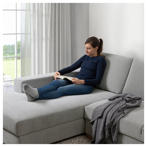 A person reads on a grey SÖRVALLEN sofa by a window, wearing a navy sweater and jeans.