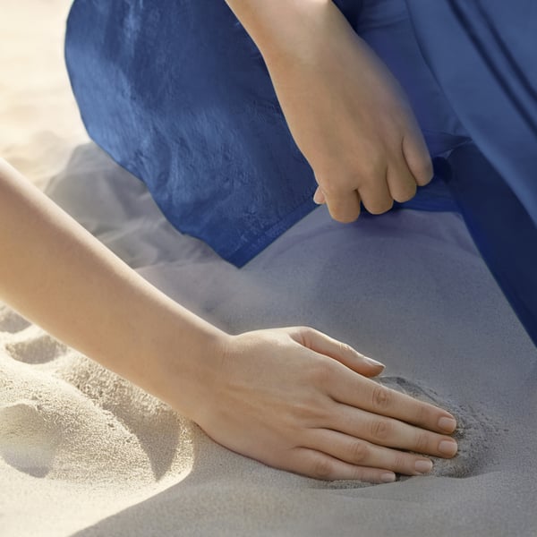 Childs hand reaches for adults hand on sandy beach under blue fabric shelter.