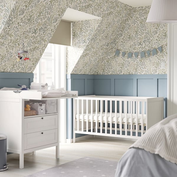 Baby nursery with SUNDVIK white cot, blue panelling, floral wallpaper, white dresser, soft toys, and polka dot rug.