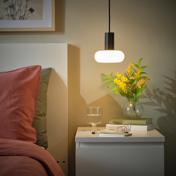 Cosy bedroom corner with SUNNEBY lamp, books, flowers, and warm pillows.