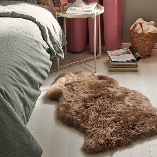 Bedroom with a green bed, pink curtains, and a brown fluffy rug on the floor. A white side table holds books and a lamp. A wicker basket is nearby.