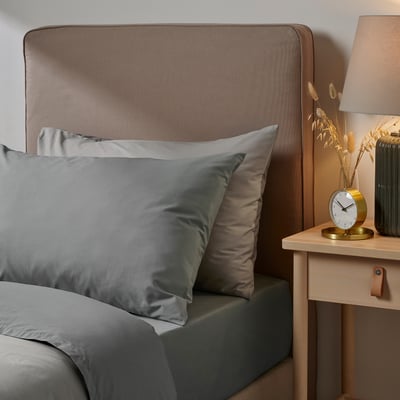 Ullvida grey pillowcase in cotton/lyocell blend by bed, rectangular shape, soft & durable. Larger than clock and lamp on wooden bedside table.