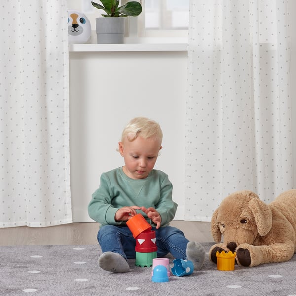 Young child playing with colourful stacking cups on the floor, surrounded by soft toys.